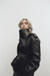 Woman with blonde hair wearing a high-collared black leather coat against a white background