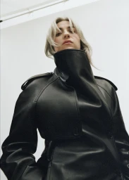 Low-angle portrait of a woman wearing a black leather coat with a high collar against a plain white background.