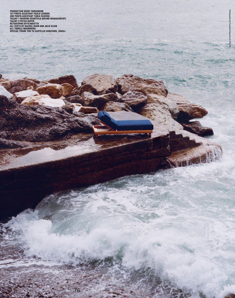 A solitary blue cushion sits atop a wooden platform on rocky steps leading to the ocean, creating a serene coastal scene.