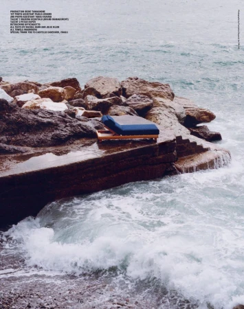 A solitary blue cushion sits atop a wooden platform on rocky steps leading to the ocean, creating a serene coastal scene.