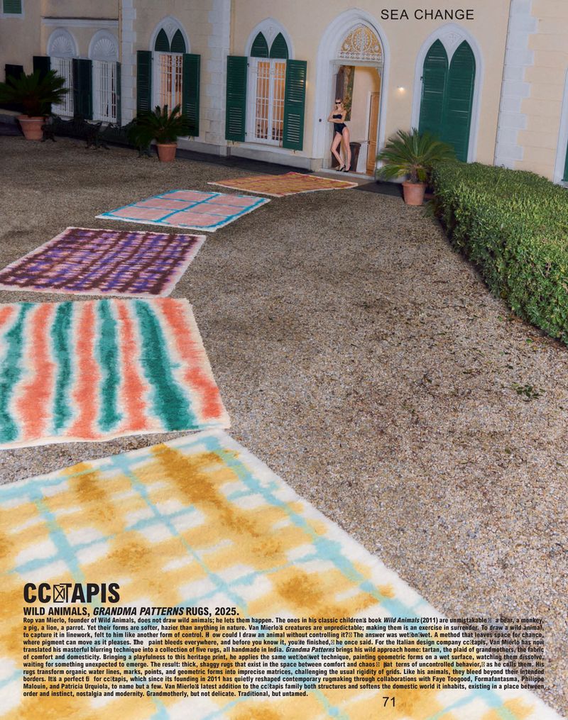 Fashion shoot features a woman in black swimsuit posing by a sunlit doorway of a pale building with colorful rugs displayed on the ground.