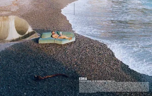 A man lying on a green outdoor sofa placed on a pebble beach next to crashing ocean waves under bright daylight.