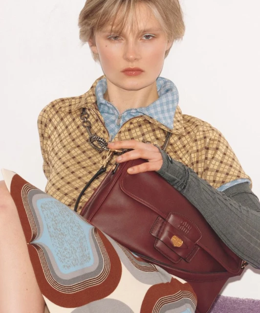 Portrait of a young woman with short blonde hair wearing a plaid shirt and a burgundy purse in a light, neutral indoor setting.
