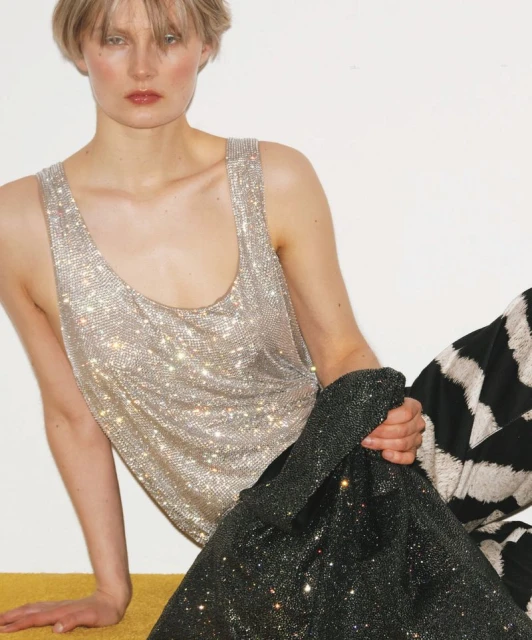 Fashion shot of a woman in a silver top and patterned pants, holding a sparkly black bag, against a plain white background.