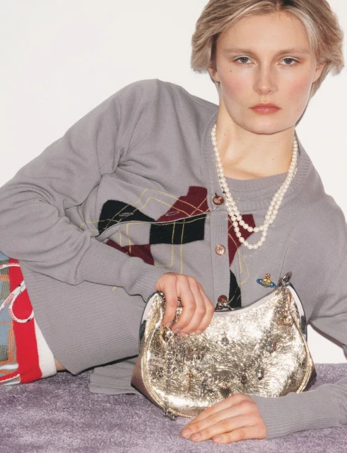 Fashion shot of a fair-skinned female model with blonde hair, wearing a grey cardigan and pearl necklace, holding a gold handbag.
