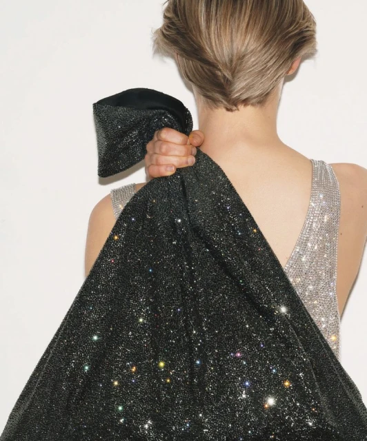 Back view of a woman with blonde hair holding a sparkly black fabric, showcasing an elegant and fashionable silver halter dress.