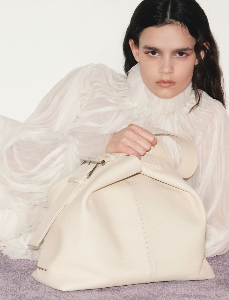 Portrait of a woman lying down in a white ruffled dress next to a beige leather satchel bag, lit by soft light.