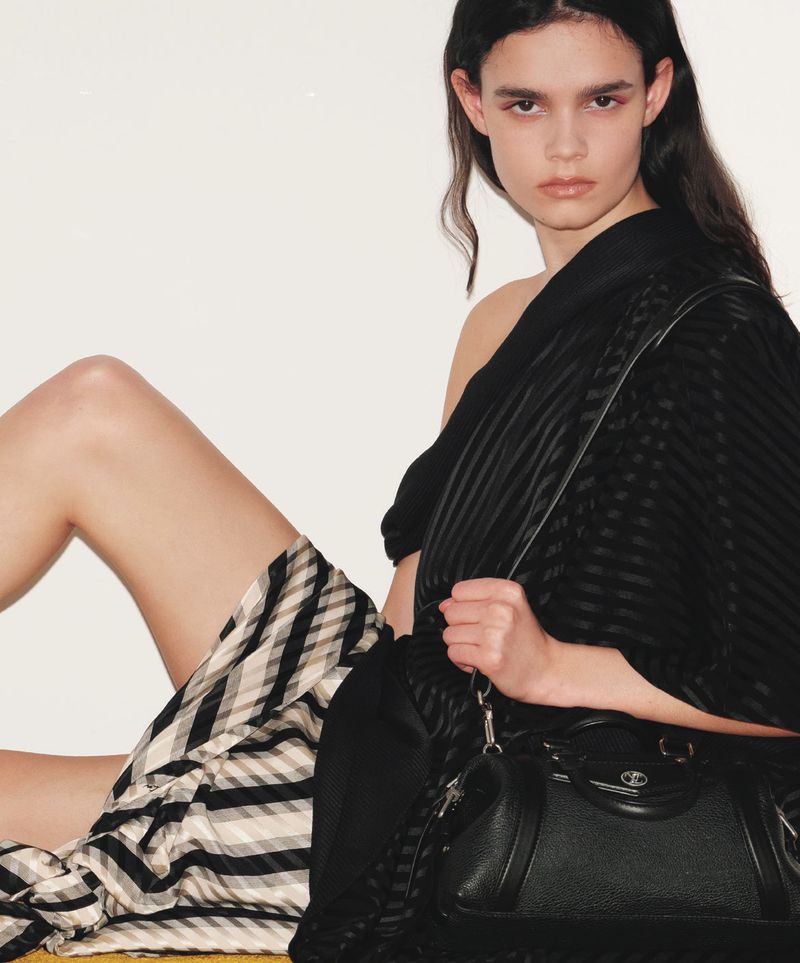 Fashion shot of a brunette model in a black top and checkered skirt sitting next to a black leather handbag in a studio setting.