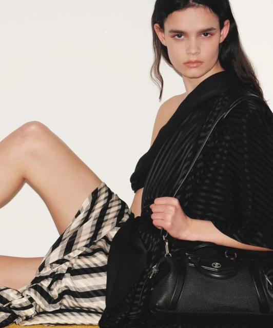 Fashion shot of a brunette model in a black top and checkered skirt sitting next to a black leather handbag in a studio setting.