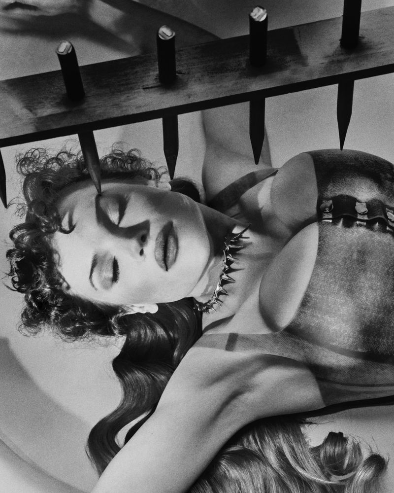 Artistic black and white image of a woman with curly hair lying below a rack of spikes, wearing a studded collar, and dramatic makeup.