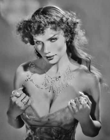 Black and white portrait of a woman with a jeweled necklace and rings, holding the edges of her translucent top, against a blurred background.