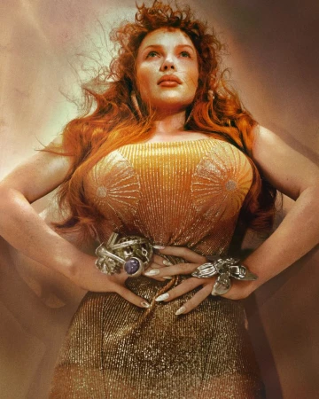 Low angle portrait of a redheaded woman wearing a shimmering gold dress and statement rings against a warm gradient background.