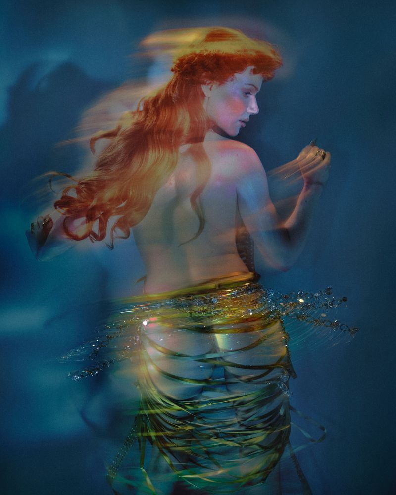 Woman with long red hair poses partially nude with green shimmering sash, motion blur, set against a soft blue background with cloudlike shadows.