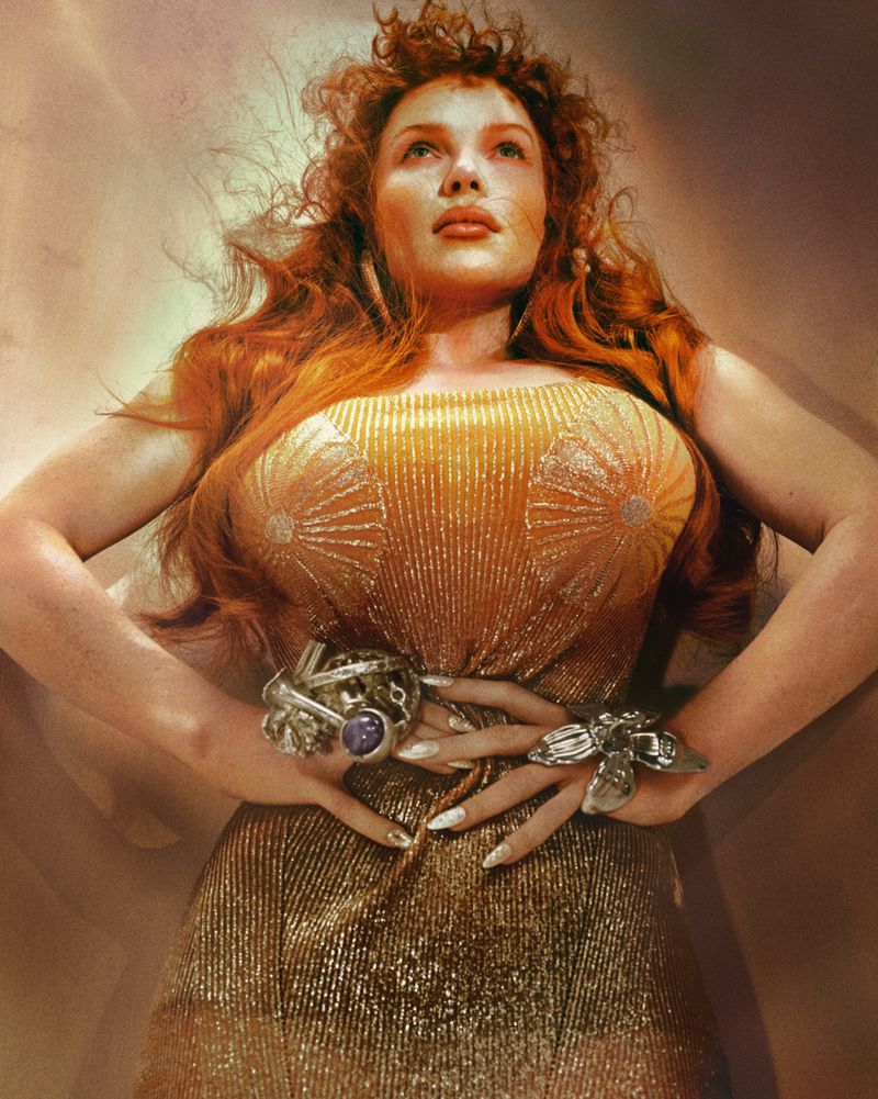 Low angle portrait of a redheaded woman wearing a shimmering gold dress and statement rings against a warm gradient background.