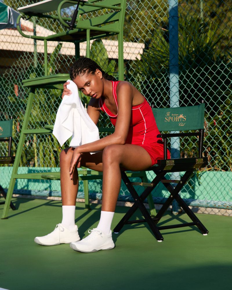 Female tennis player seated on court in red uniform, wiping sweat with towel, next to tennis chair. Outdoor setting, bright sunlight.