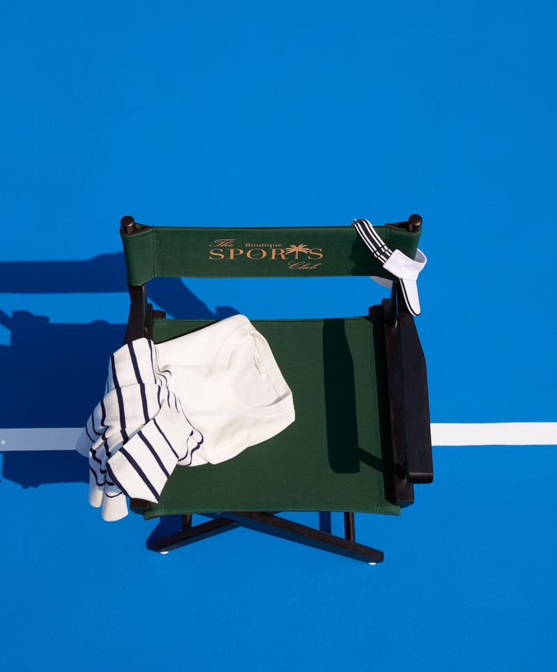 Overhead shot of an empty green canvas director's chair on a blue tennis court, with clothing and accessories arranged on the seat.