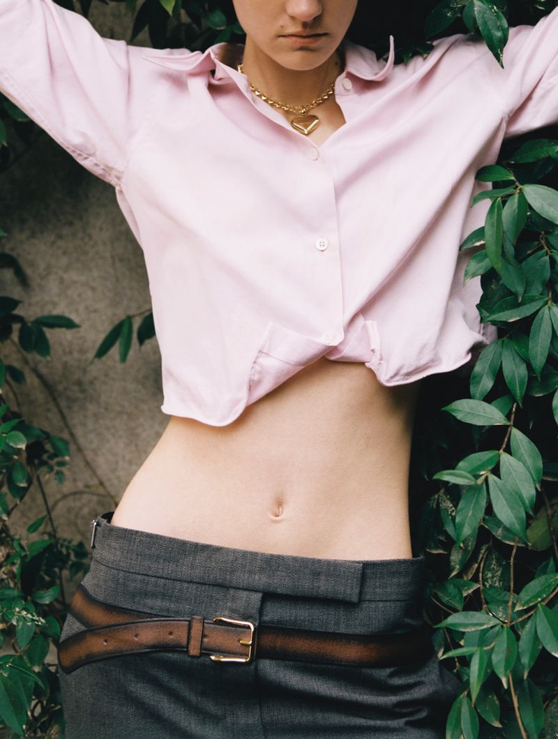 Mid-section of a young adult woman in a pink shirt and grey pants posing outdoors near a green leafy wall with a gold heart necklace.