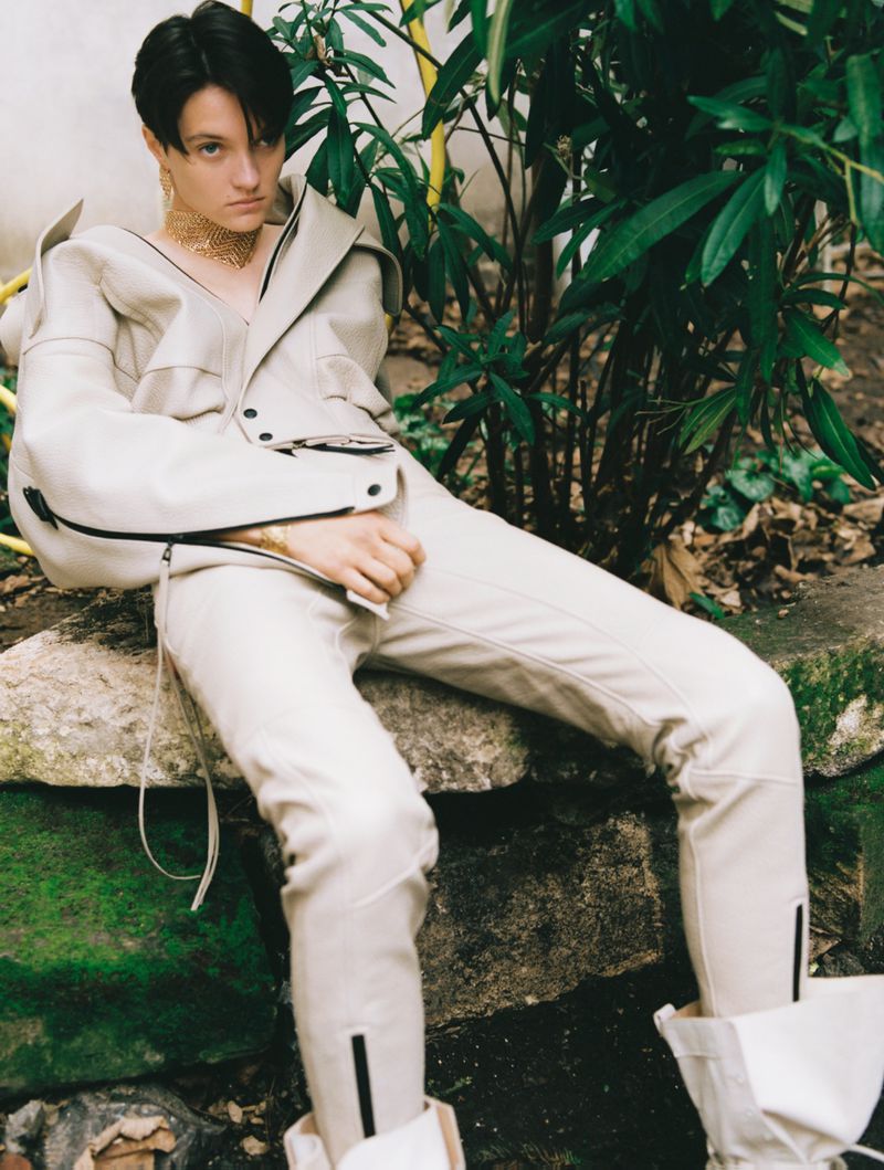 Person wearing off-white outfit posing in an outdoor setting surrounded by plants and a stone structure, captured in a medium shot.