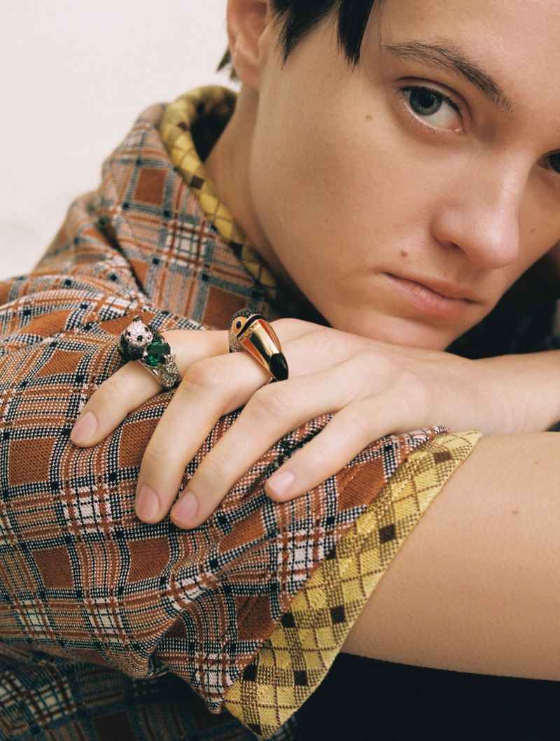Person with fair skin rests their chin on their crossed hands, adorned with rings; wearing a plaid shirt against a white background.