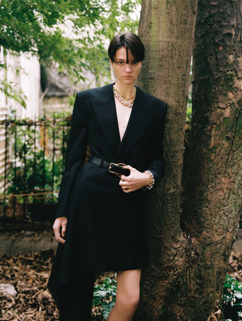Woman poses next to a tree wearing a black belted dress, gold necklace, and bracelet; outdoor fashion shoot.