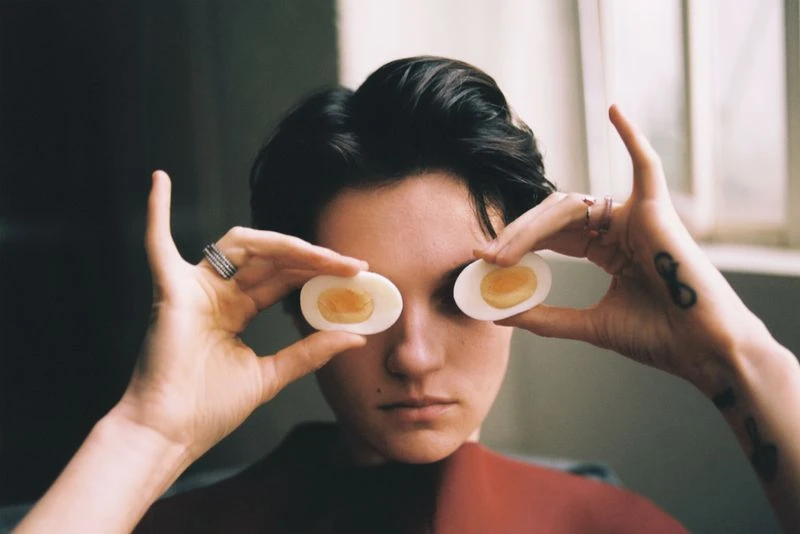 A woman holds two hard-boiled egg halves up to her eyes in an indoor portrait with shallow depth of field and natural lighting.