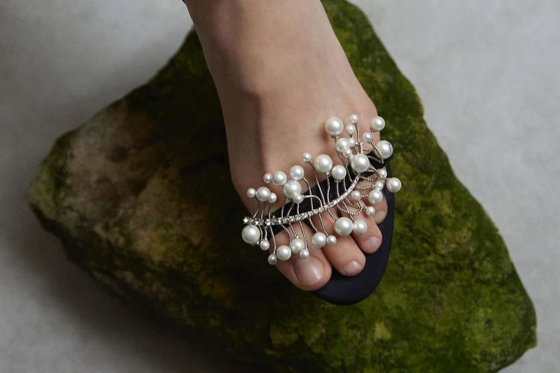 A foot wears a black sandal with a pearl and crystal embellishment atop a stone surface. The shot is a close-up with soft lighting.