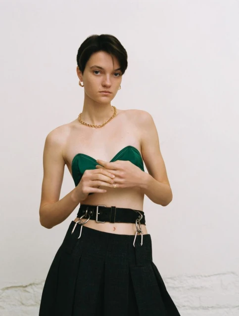 A young woman with short dark hair poses indoors wearing a green strapless top and a black skirt with gold jewelry against a white wall.