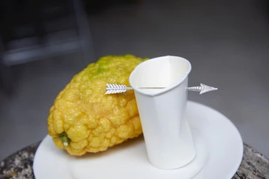 A textured yellow citrus fruit and a white paper cup pierced by a decorative arrow-shaped brooch on a white plate.