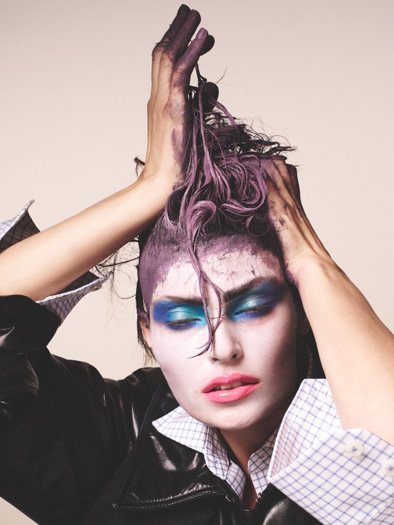 Close-up portrait of an adult female with purple hair and bold makeup, wearing a black jacket and a patterned shirt, set against a neutral background.