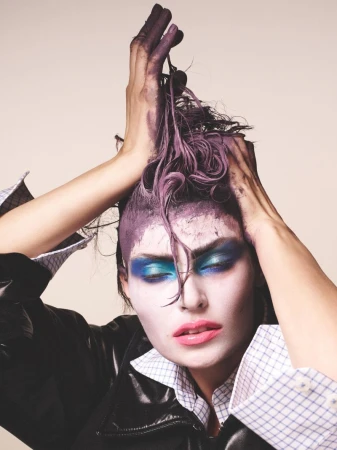 Close-up portrait of an adult female with purple hair and bold makeup, wearing a black jacket and a patterned shirt, set against a neutral background.