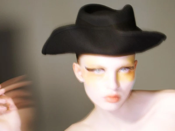 Model wearing a black wide-brimmed hat with yellow eyeshadow and motion-blurred hand against a neutral background