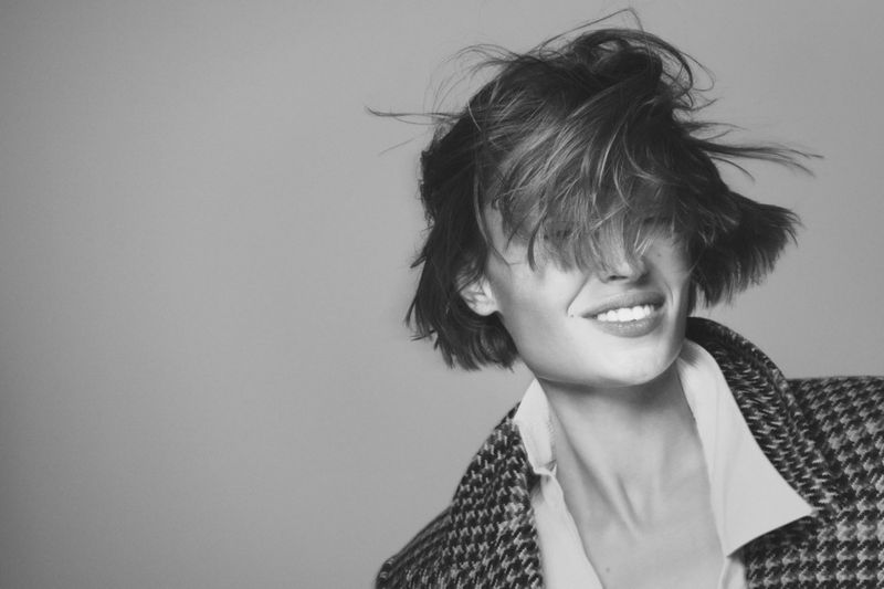 Portrait of smiling woman with tousled hair wearing a patterned coat against a neutral grey background