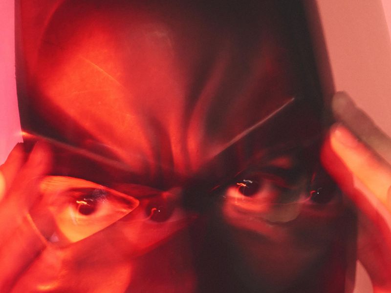 Close-up of a person's face partially obscured by their fingers and a red filter, with only their eyes visible against a light pink background.