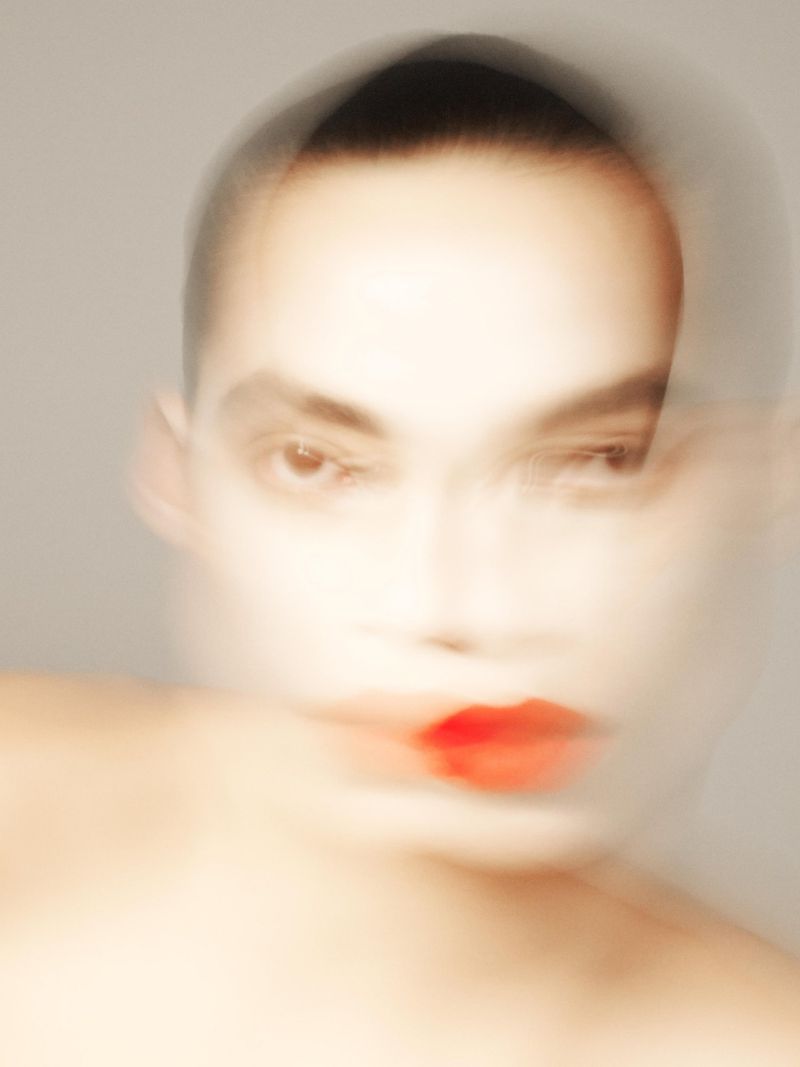 Close-up portrait of a blurry human face with bright red lipstick and short dark hair against a muted grey background.