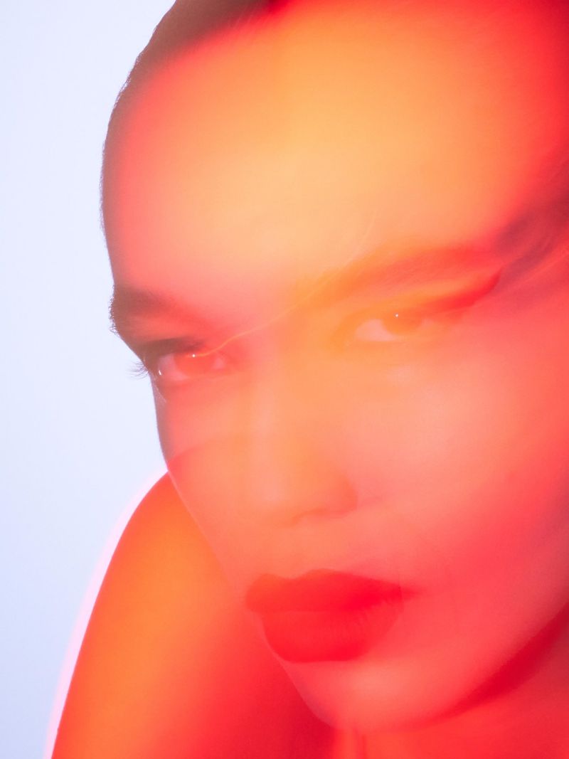 Portrait of a woman with shaved head, close-up, dramatically lit with red and orange hues against a pale blue background.