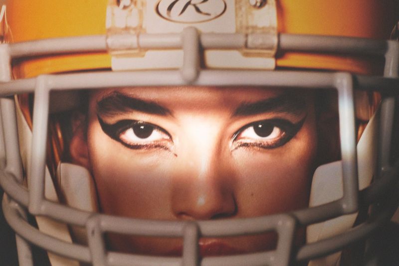 Close-up of a person's face with dramatic eyeliner, partially obscured by a football helmet, with a light shining across the face.