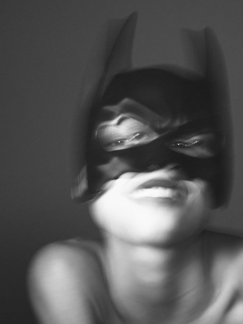 Blurry black and white portrait of a person wearing a black mask with pointed ears and a bare upper body in front of a gray background.