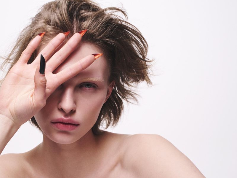 Close-up beauty portrait of a model with short tousled hair holding a hand with pointed orange and black nails in front of her face.
