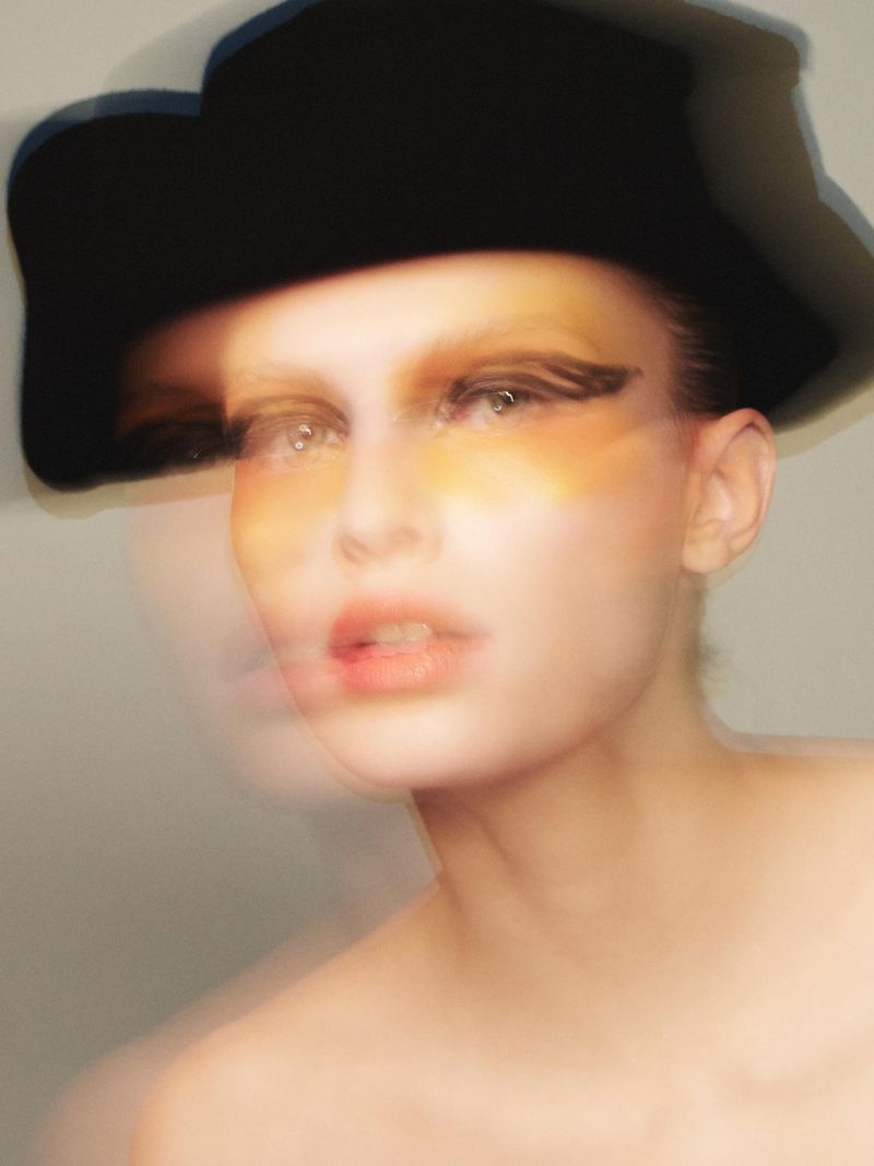 Headshot of a fair-skinned woman wearing a black hat and heavy eye makeup with yellow eyeshadow and black eyeliner. Soft-focus.