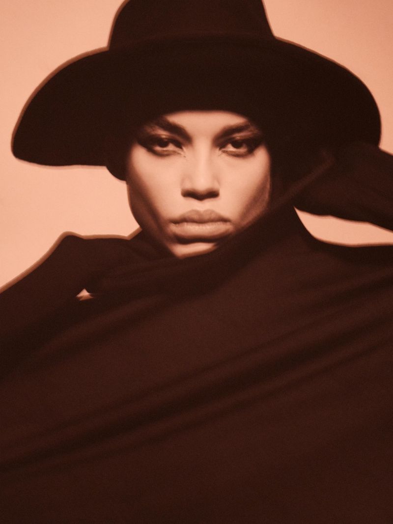 Elegant portrait of a woman with dark hair and hat, framed by dark fabric. The image is lit with warm tones in a minimalist studio setting.