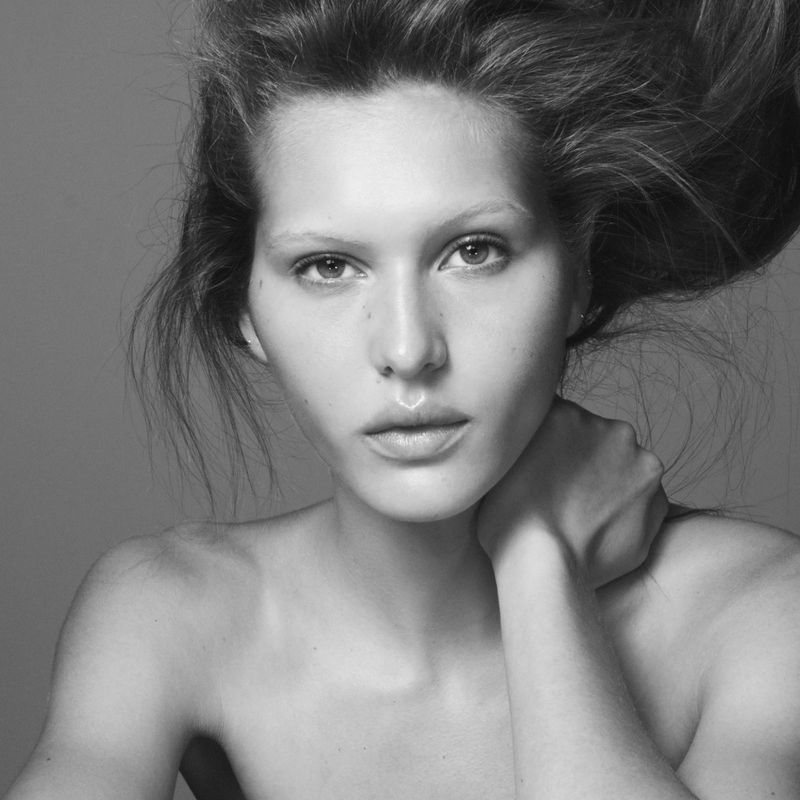 Black and white portrait of an adult woman with loose hair posing with a hand on her neck against a neutral grey backdrop in studio.
