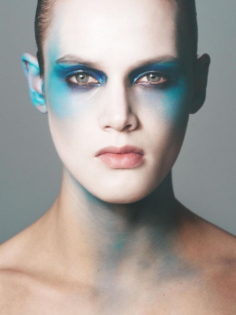 Close-up portrait of a young adult woman with bold blue makeup around her eyes, temples, and neck against a soft grey background.