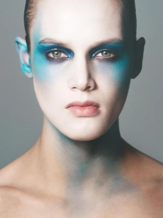 Close-up portrait of a young adult woman with bold blue makeup around her eyes, temples, and neck against a soft grey background.