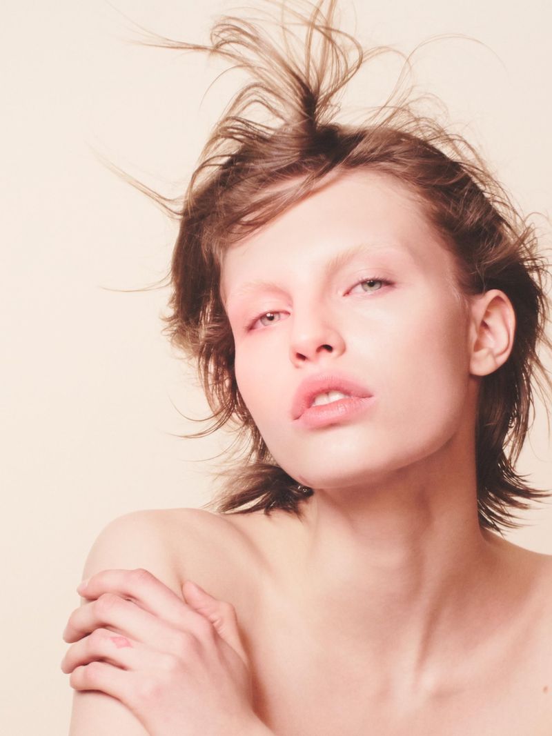 Headshot of a young adult woman with tousled short hair and bare shoulders against a light peach-toned background in soft studio lighting.