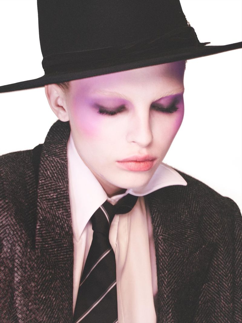 Model wearing a black hat, tweed blazer, white shirt, and tie with purple eye makeup against a white backdrop