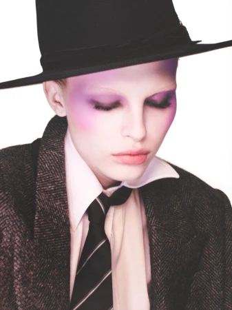 Model wearing a black hat, tweed blazer, white shirt, and tie with purple eye makeup against a white backdrop