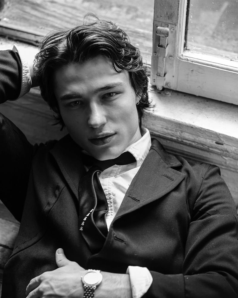 Black-and-white portrait of a young adult male with wavy dark hair, wearing a dark blazer and light shirt, lying near a window.