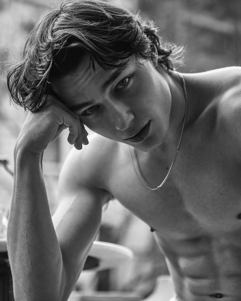Black and white portrait of a shirtless young adult male with dark wavy hair, a chain necklace, and a contemplative expression.