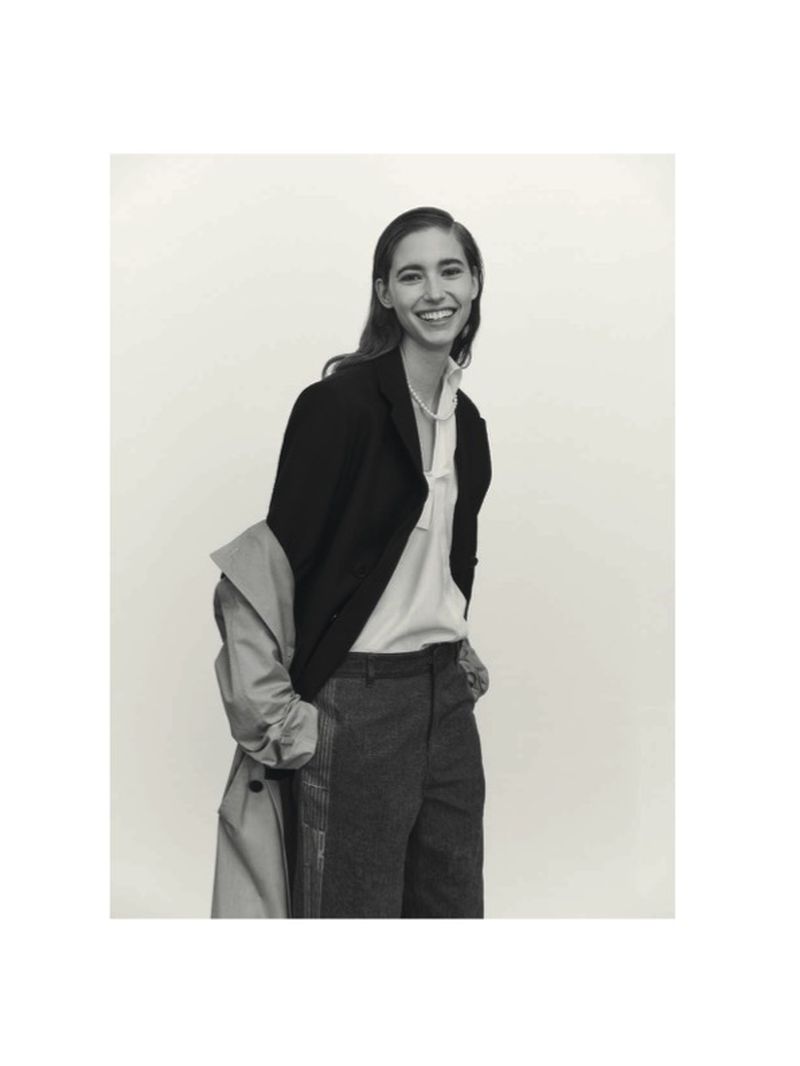 Smiling woman in black blazer and grey trousers standing indoors with an unbuttoned beige trench coat on white backdrop.