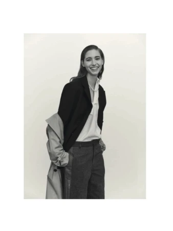 Smiling woman in black blazer and grey trousers standing indoors with an unbuttoned beige trench coat on white backdrop.